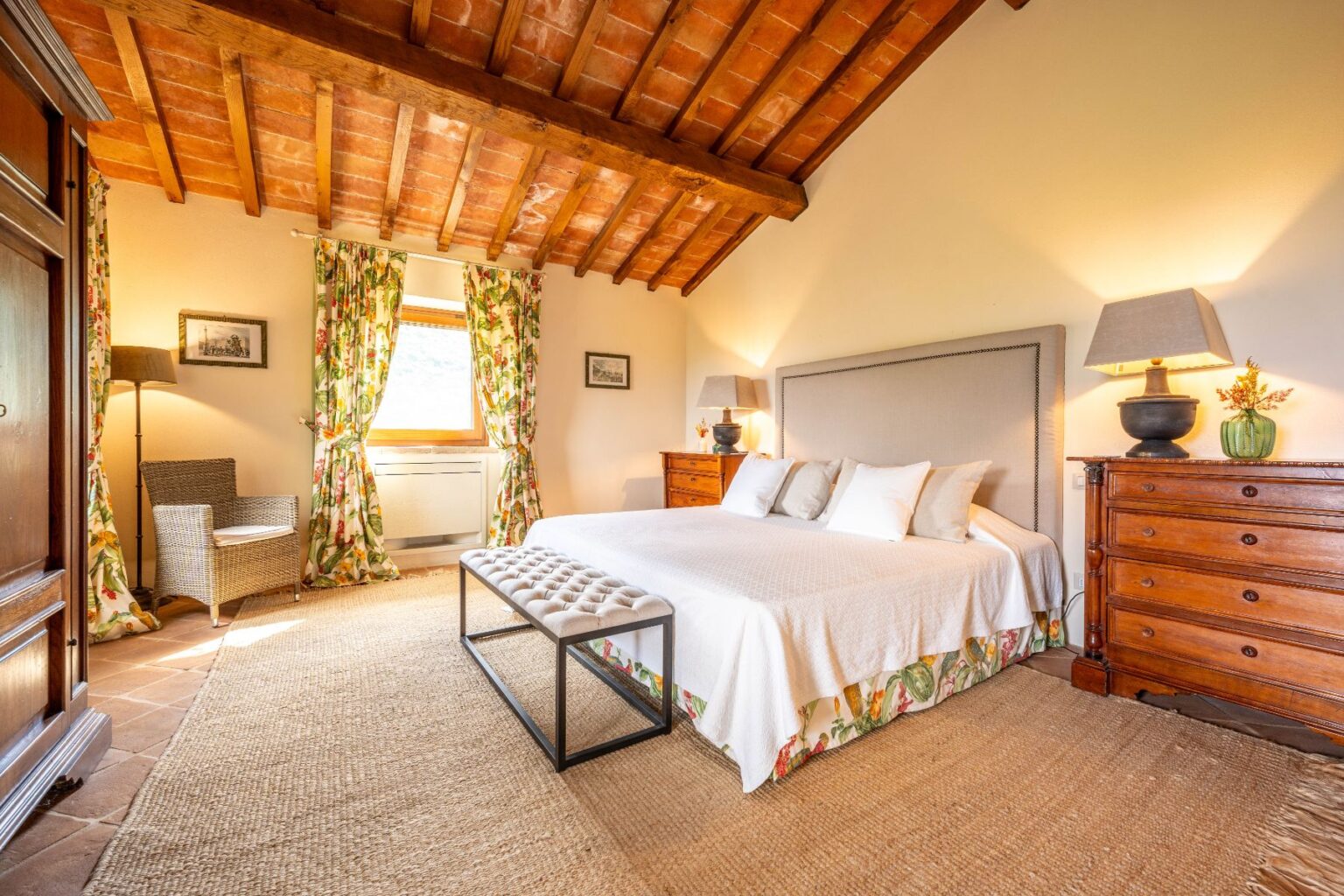 Cozy bedroom featuring a king-sized bed with white linens, decorative pillows, and a stylish headboard. The room has wooden beams, floral curtains, a woven rug, and a bedside table with a lamp. A comfortable armchair and a wooden dresser add to the inviting atmosphere. Natural light streams in through the window, enhancing the warm ambiance. | Only on SopranoVillas