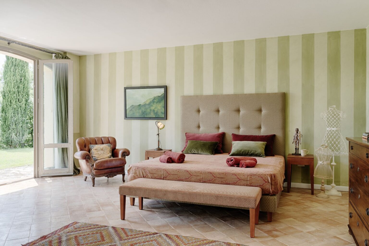 Spacious and stylish bedroom featuring a king-size bed with plush pillows, a cozy leather armchair, and elegant striped walls. The room includes a decorative bench at the foot of the bed, a wooden nightstand with a lamp, and a view of the garden through large glass doors. Ideal for a relaxing retreat. | Only on SopranoVillas