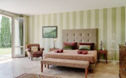 Spacious and stylish bedroom featuring a king-size bed with plush pillows, a cozy leather armchair, and elegant striped walls. The room includes a decorative bench at the foot of the bed, a wooden nightstand with a lamp, and a view of the garden through large glass doors. Ideal for a relaxing retreat.