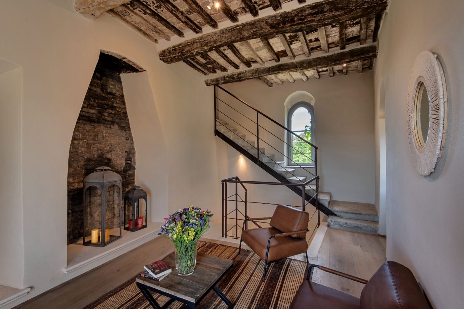 Modern living room with rustic wooden beams, featuring a stylish staircase, comfortable leather chairs, a coffee table with books, and decorative lanterns. Fresh flowers in a vase add a pop of color to the serene interior. | Only on SopranoVillas