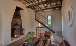 Modern living room with rustic wooden beams, featuring a stylish staircase, comfortable leather chairs, a coffee table with books, and decorative lanterns. Fresh flowers in a vase add a pop of color to the serene interior.