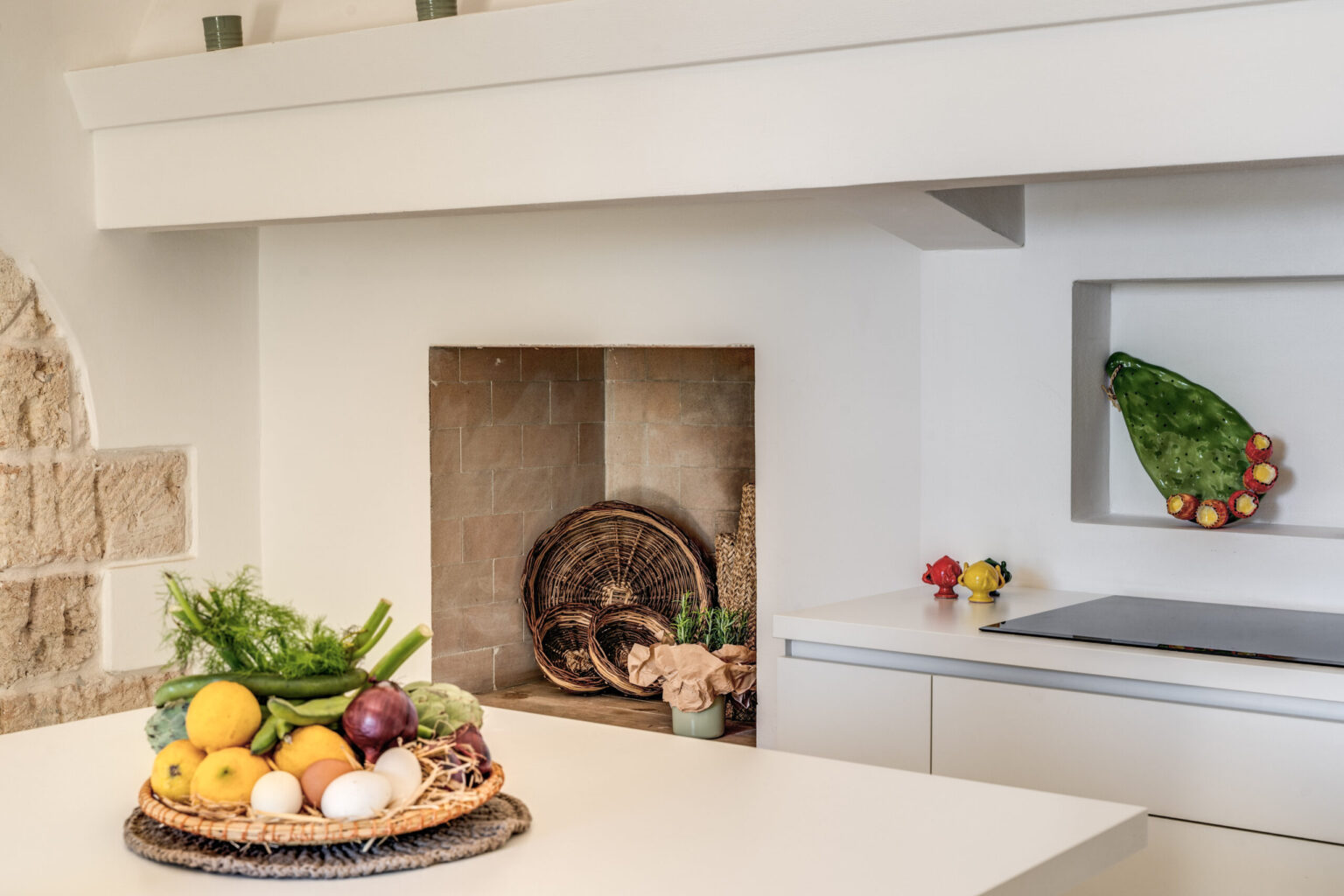 Modern kitchen interior featuring a stylish fruit basket with various fruits, green vegetables, and a minimalist design. The kitchen includes a sleek countertop, a built-in stove, and a decorative wall niche with additional fruits, emphasizing a clean and inviting atmosphere. | Only on SopranoVillas