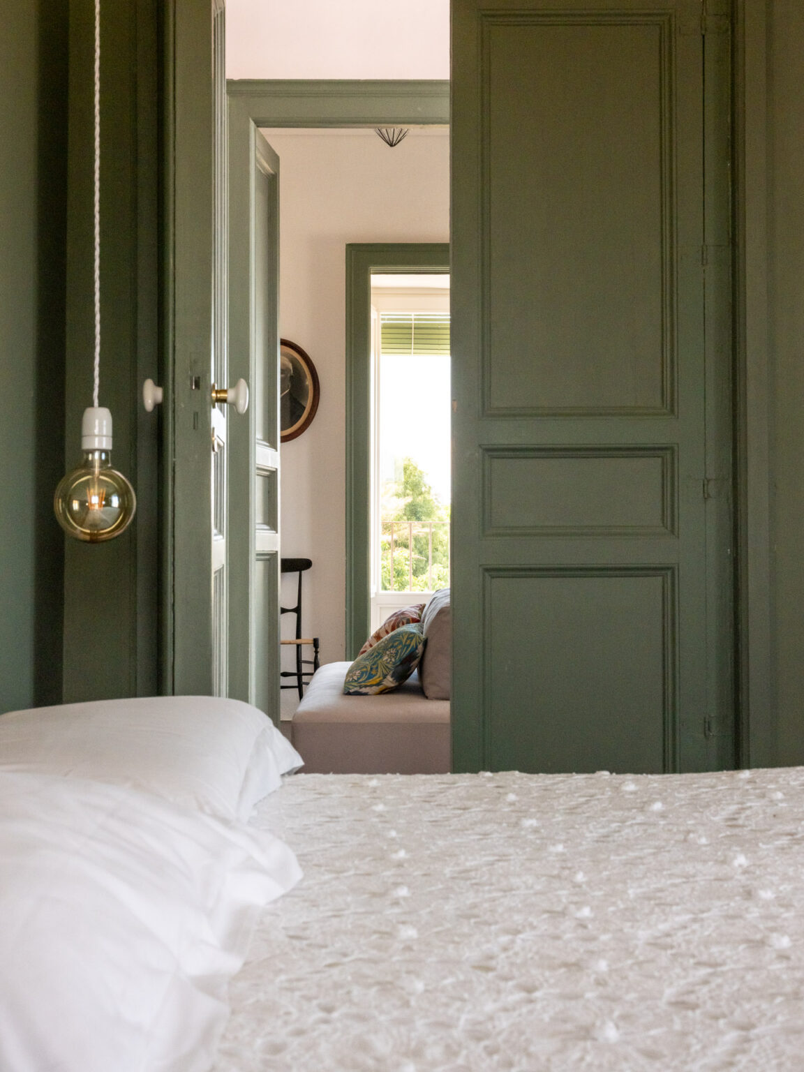 Cozy bedroom view through open green doors, featuring a white textured bedspread and a decorative pillow on a nearby sofa, with natural light streaming in from a window. | Only on SopranoVillas