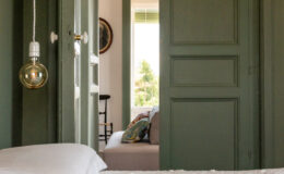 Cozy bedroom view through open green doors, featuring a white textured bedspread and a decorative pillow on a nearby sofa, with natural light streaming in from a window.