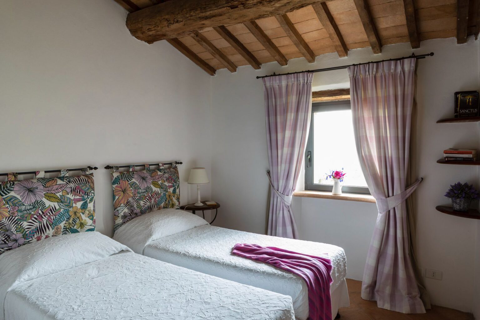 Cozy bedroom featuring two twin beds with floral headboards, soft white bedding, and a purple throw. Natural light streams in through a window adorned with plaid curtains, complemented by a small shelf with decorative items and a vase of flowers. Rustic wooden beams enhance the room's charm. | Only on SopranoVillas