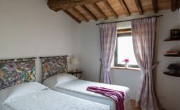 Cozy bedroom featuring two twin beds with floral headboards, soft white bedding, and a purple throw. Natural light streams in through a window adorned with plaid curtains, complemented by a small shelf with decorative items and a vase of flowers. Rustic wooden beams enhance the room's charm.