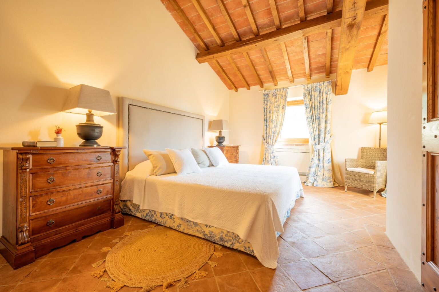 Cozy bedroom featuring a large double bed with white bedding, wooden furniture, and a rustic ceiling. The room includes a bedside table with a lamp, a woven rug, and floral curtains that frame the window, creating a warm and inviting atmosphere. | Only on SopranoVillas