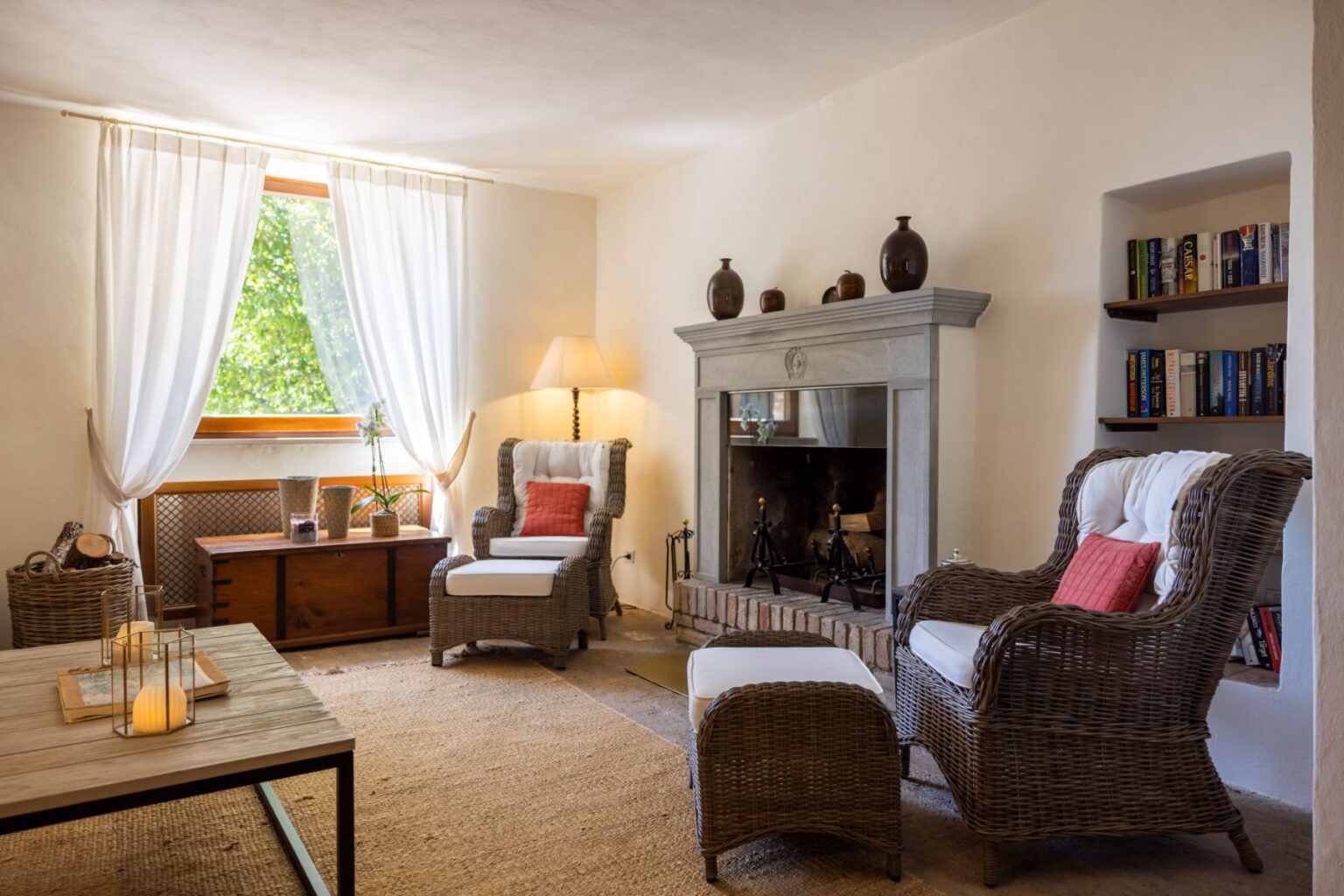 Cozy living room featuring wicker chairs with white cushions, a stone fireplace, and a wooden coffee table. Natural light enters through sheer curtains, highlighting decorative elements like candles and bookshelves. | Only on SopranoVillas