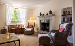 Cozy living room featuring wicker chairs with white cushions, a stone fireplace, and a wooden coffee table. Natural light enters through sheer curtains, highlighting decorative elements like candles and bookshelves.
