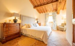 Cozy bedroom featuring a large double bed with white bedding, wooden furniture, and a rustic ceiling. The room includes a bedside table with a lamp, a woven rug, and floral curtains that frame the window, creating a warm and inviting atmosphere.