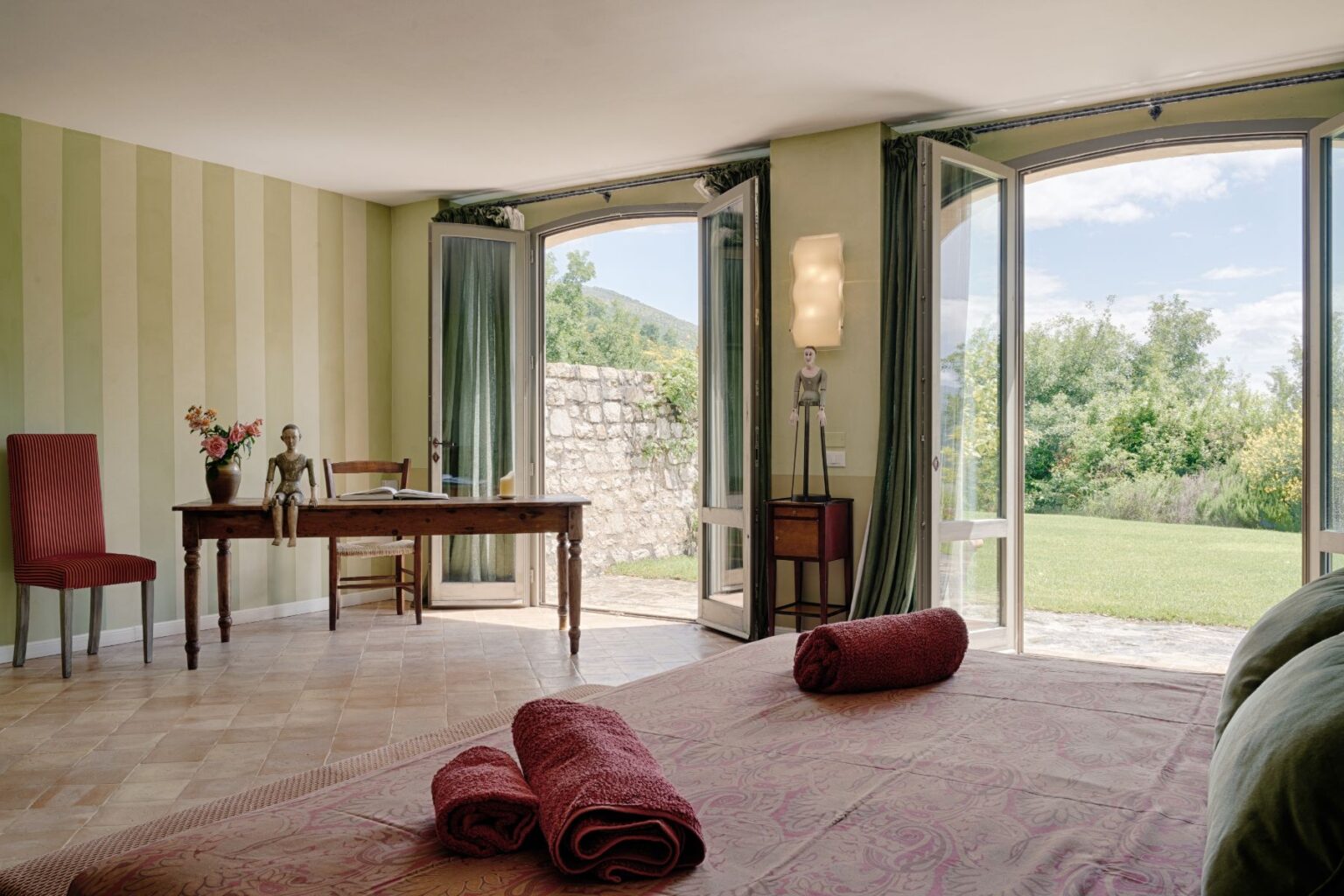 Bright and airy bedroom featuring large open windows with scenic views, a wooden desk with a decorative mannequin, a red striped chair, and a bed adorned with rolled towels. The room is decorated with soft green striped walls and has a cozy, inviting atmosphere. | Only on SopranoVillas