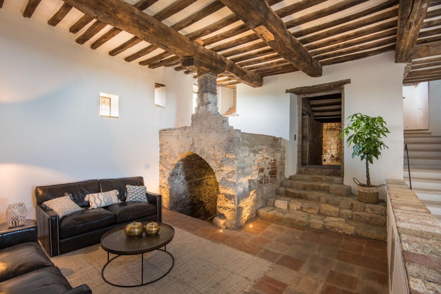 Cozy living room featuring a leather sofa with decorative pillows, a round coffee table with metallic accents, and a rustic stone fireplace. Exposed wooden beams and a potted plant add character to the space, while a staircase leads to the upper level. | Only on SopranoVillas
