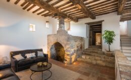 Cozy living room featuring a leather sofa with decorative pillows, a round coffee table with metallic accents, and a rustic stone fireplace. Exposed wooden beams and a potted plant add character to the space, while a staircase leads to the upper level.