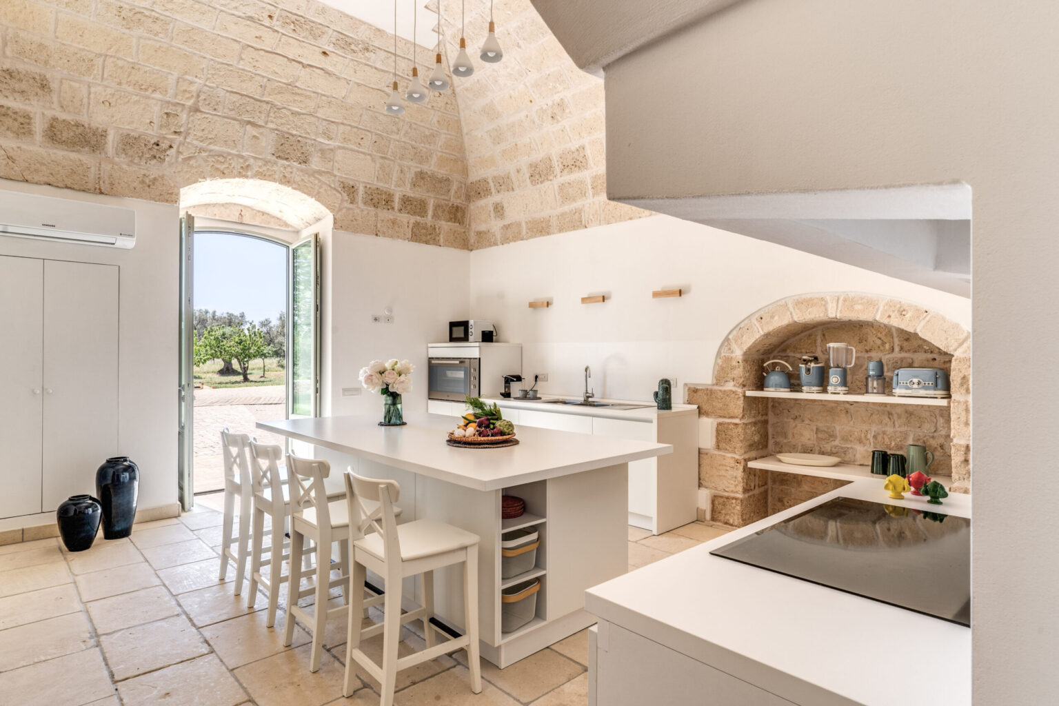 Modern open-concept kitchen featuring a large white island, stone walls, and natural light streaming through arched windows, with stylish bar stools and a cozy dining area. | Only on SopranoVillas