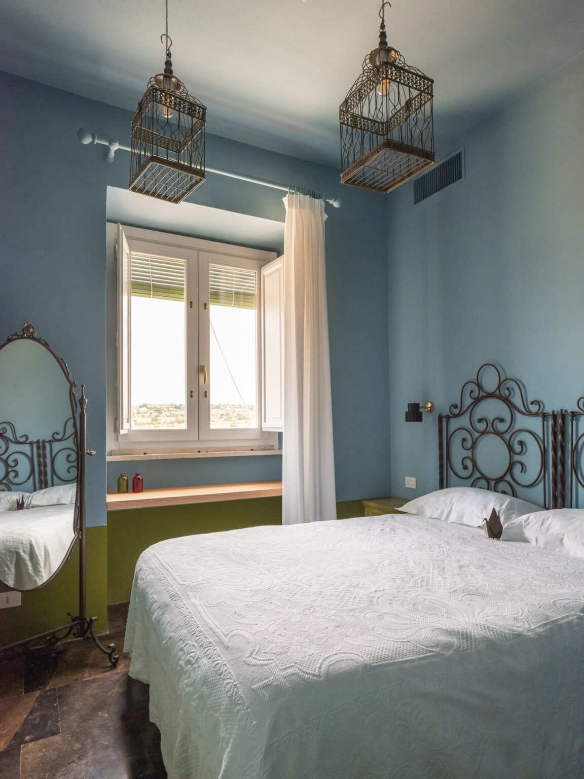 Cozy bedroom featuring a white quilted bedspread, decorative birdcage light fixtures, and a vintage mirror. Bright blue walls and a window with sheer curtains create a welcoming atmosphere, complemented by a minimalist wooden shelf. Ideal for relaxation and comfort. | Only on SopranoVillas