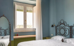 Cozy bedroom featuring a white quilted bedspread, decorative birdcage light fixtures, and a vintage mirror. Bright blue walls and a window with sheer curtains create a welcoming atmosphere, complemented by a minimalist wooden shelf. Ideal for relaxation and comfort.