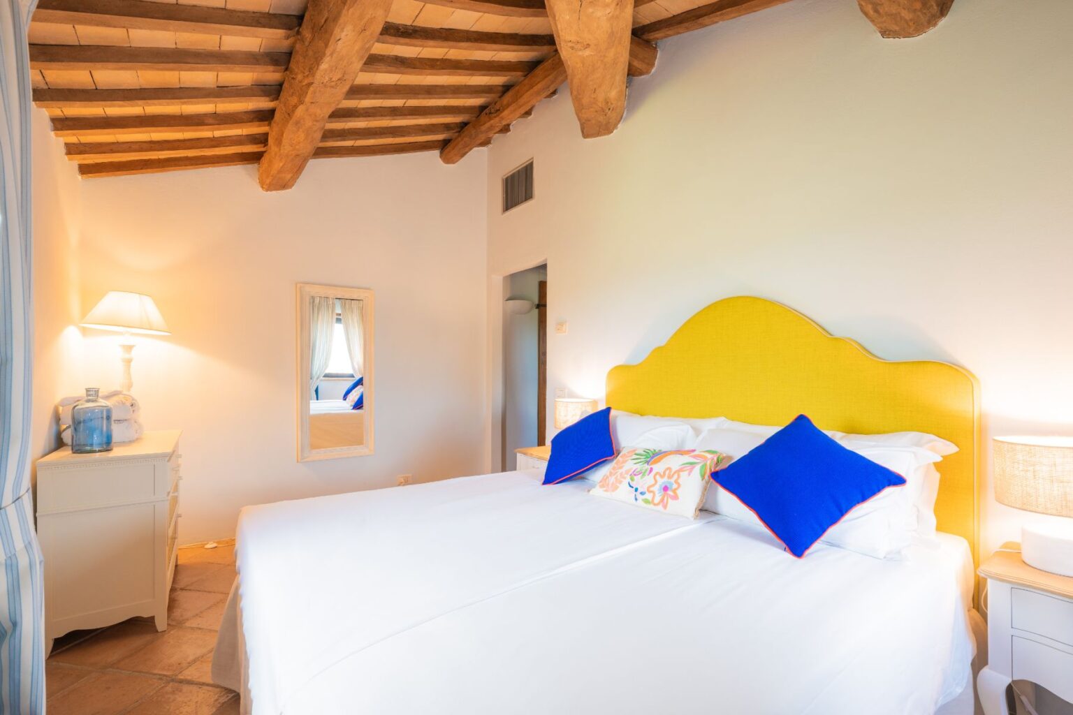 Cozy bedroom featuring a yellow upholstered headboard, white bedding, and colorful accent pillows. The room includes a bedside lamp, a mirror, and rustic wooden beams on the ceiling, creating a warm and inviting atmosphere. | Only on SopranoVillas