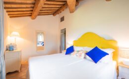 Cozy bedroom featuring a yellow upholstered headboard, white bedding, and colorful accent pillows. The room includes a bedside lamp, a mirror, and rustic wooden beams on the ceiling, creating a warm and inviting atmosphere.