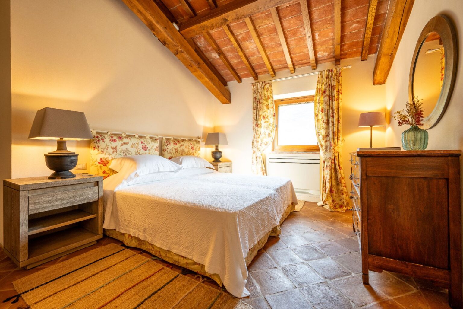 Cozy bedroom featuring a queen-sized bed with a floral headboard, wooden ceiling beams, and soft lighting. The room includes bedside tables with lamps, a vintage dresser, and large windows adorned with floral curtains, creating a warm and inviting atmosphere. Ideal for relaxation and comfort. | Only on SopranoVillas