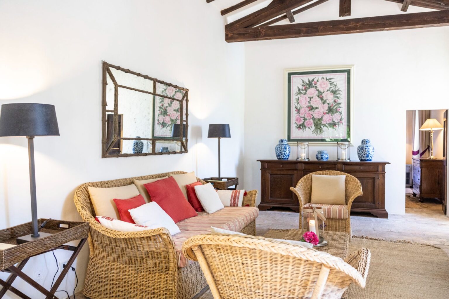 Cozy living room featuring a wicker sofa with colorful cushions, two matching armchairs, and a wooden coffee table. Decorated with a large floral artwork, a decorative mirror, and blue ceramic vases. Soft lighting from a floor lamp enhances the inviting atmosphere. | Only on SopranoVillas