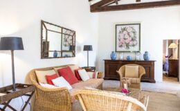 Cozy living room featuring a wicker sofa with colorful cushions, two matching armchairs, and a wooden coffee table. Decorated with a large floral artwork, a decorative mirror, and blue ceramic vases. Soft lighting from a floor lamp enhances the inviting atmosphere.