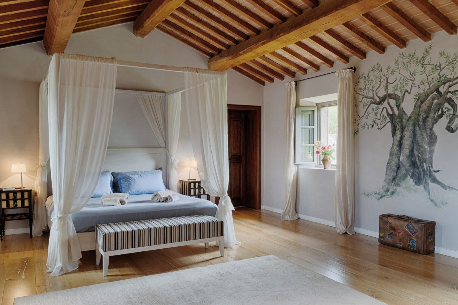 Spacious and elegantly designed bedroom featuring a four-poster bed with sheer drapes, wooden beams, and a decorative mural of an olive tree. The room includes a cozy striped bench, bedside tables with lamps, and a window adorned with flowers, creating a serene and inviting atmosphere. | Only on SopranoVillas