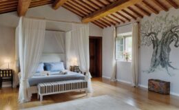 Spacious and elegantly designed bedroom featuring a four-poster bed with sheer drapes, wooden beams, and a decorative mural of an olive tree. The room includes a cozy striped bench, bedside tables with lamps, and a window adorned with flowers, creating a serene and inviting atmosphere.