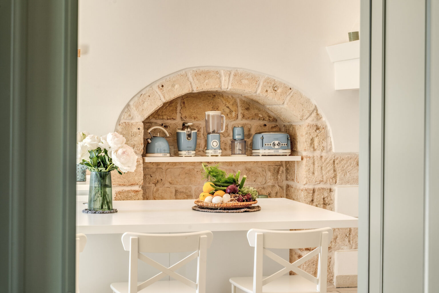 Bright and inviting kitchen featuring a stone archway, white dining table with a fruit platter, and decorative blue kitchenware on shelves. Fresh flowers in a vase add a touch of elegance to the space. | Only on SopranoVillas