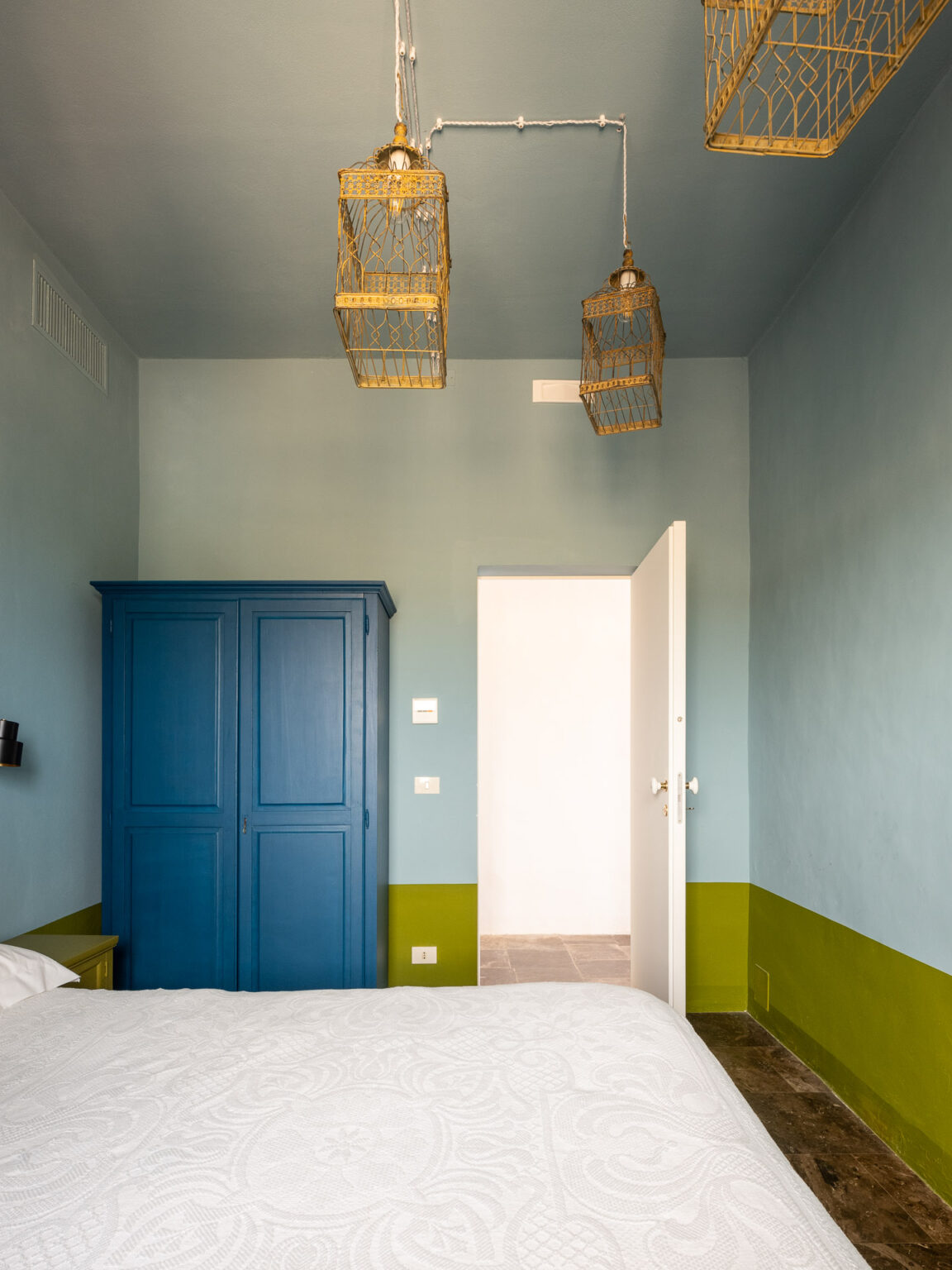 Brightly lit bedroom featuring a white bedspread, a blue wardrobe, and unique gold birdcage light fixtures hanging from the ceiling. The walls are painted in soft blue and green tones, creating a fresh and inviting atmosphere. A white door leads to another room, enhancing the spacious feel of the design. | Only on SopranoVillas