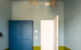 Brightly lit bedroom featuring a white bedspread, a blue wardrobe, and unique gold birdcage light fixtures hanging from the ceiling. The walls are painted in soft blue and green tones, creating a fresh and inviting atmosphere. A white door leads to another room, enhancing the spacious feel of the design.