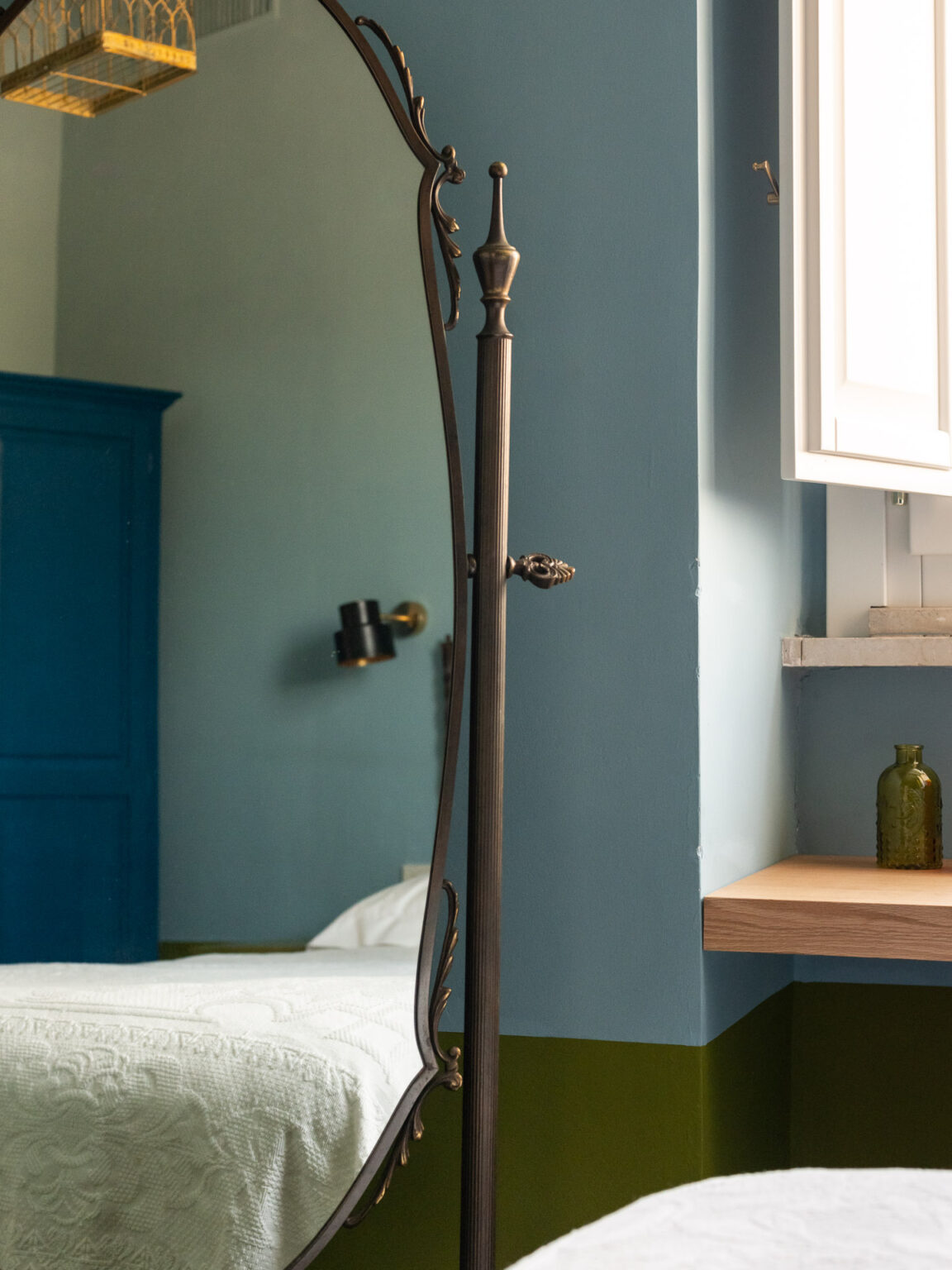 Full-length vintage mirror in a stylish bedroom with blue and green walls, featuring a wooden shelf and decorative items, complemented by a cozy bed with a white quilt. | Only on SopranoVillas