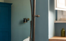 Full-length vintage mirror in a stylish bedroom with blue and green walls, featuring a wooden shelf and decorative items, complemented by a cozy bed with a white quilt.