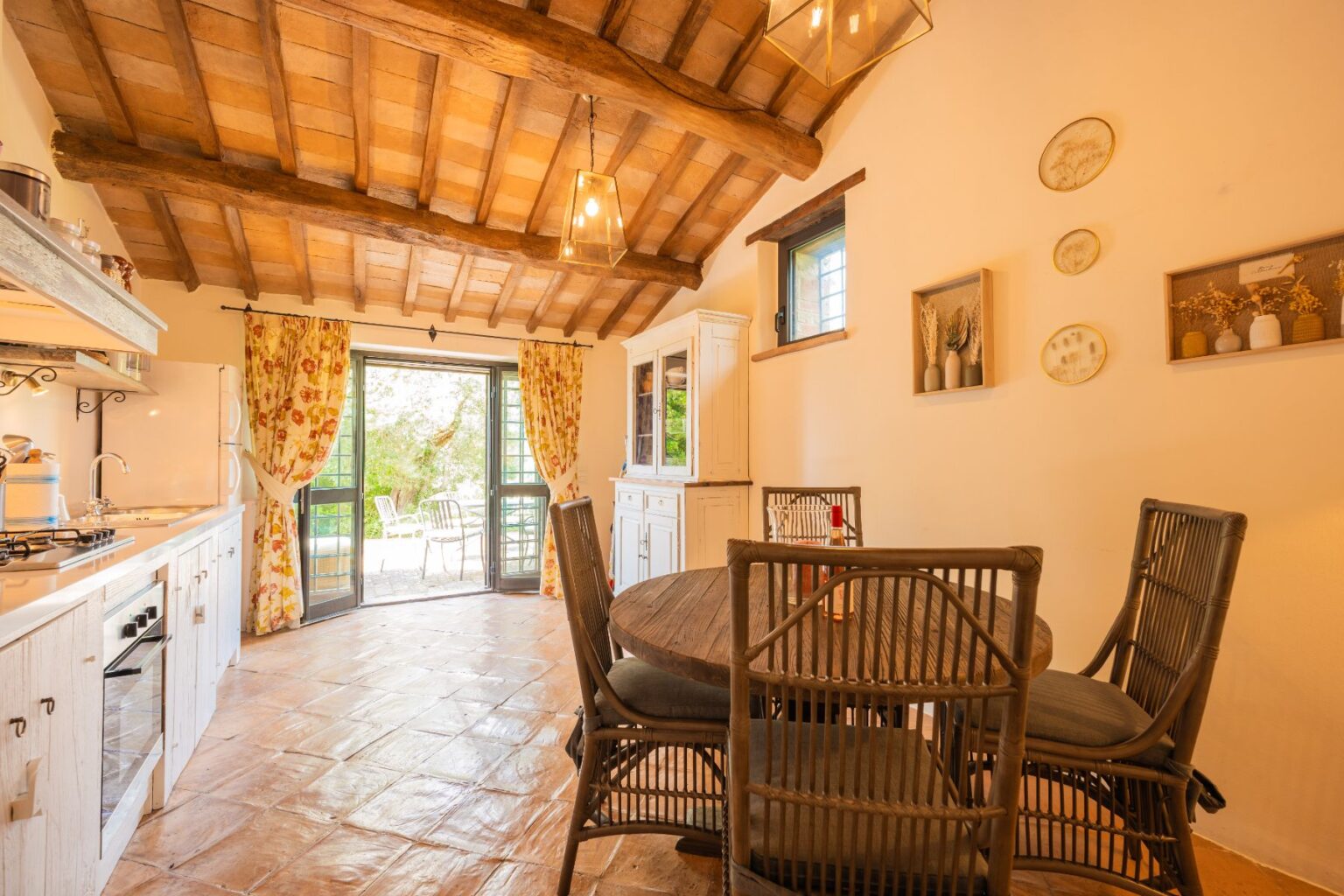 Cozy rustic kitchen and dining area featuring wooden beams, a round table with wicker chairs, and floral curtains. Large open doors lead to an outdoor patio surrounded by greenery, enhancing the inviting atmosphere. | Only on SopranoVillas