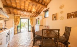 Cozy rustic kitchen and dining area featuring wooden beams, a round table with wicker chairs, and floral curtains. Large open doors lead to an outdoor patio surrounded by greenery, enhancing the inviting atmosphere.