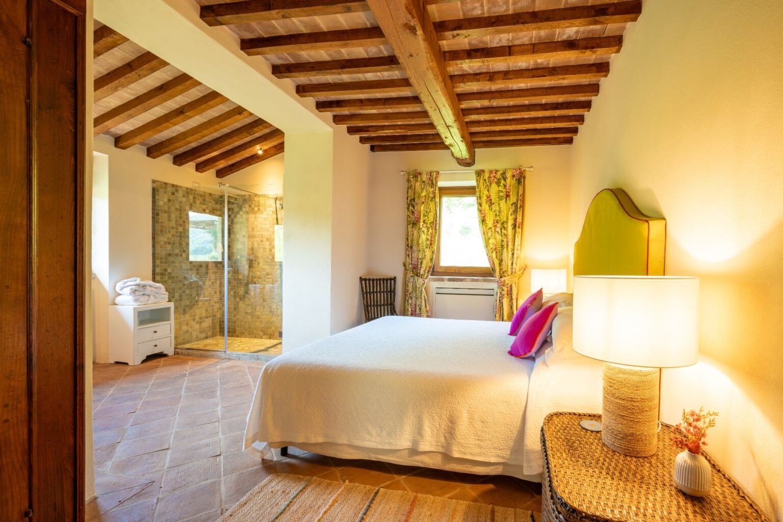 Cozy bedroom featuring a queen-sized bed with colorful accent pillows, warm wooden beams, and a modern en-suite bathroom with a glass shower. The room is adorned with floral curtains and a stylish lamp, creating a welcoming atmosphere. | Only on SopranoVillas