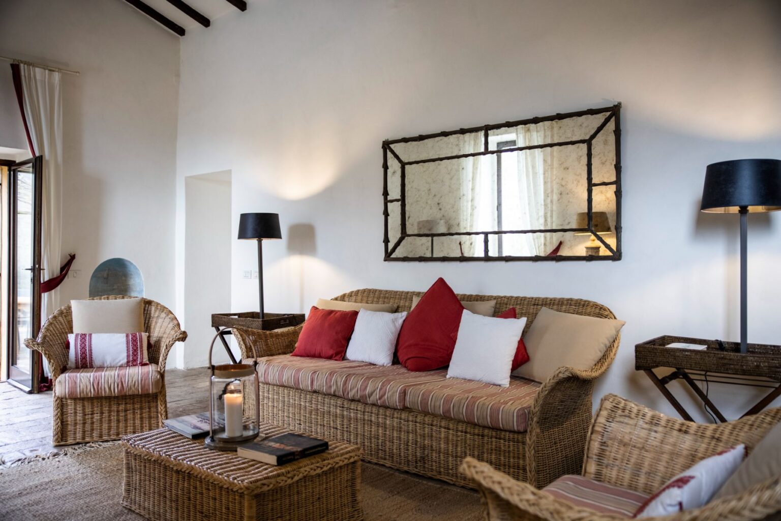 Cozy living room featuring a woven rattan sofa adorned with red and white cushions, complemented by matching armchairs. A stylish coffee table with books and a candle adds warmth, while a rustic mirror reflects natural light from the window. The space is enhanced by a modern floor lamp and neutral-toned decor, creating a welcoming atmosphere. | Only on SopranoVillas
