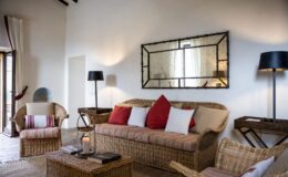Cozy living room featuring a woven rattan sofa adorned with red and white cushions, complemented by matching armchairs. A stylish coffee table with books and a candle adds warmth, while a rustic mirror reflects natural light from the window. The space is enhanced by a modern floor lamp and neutral-toned decor, creating a welcoming atmosphere.