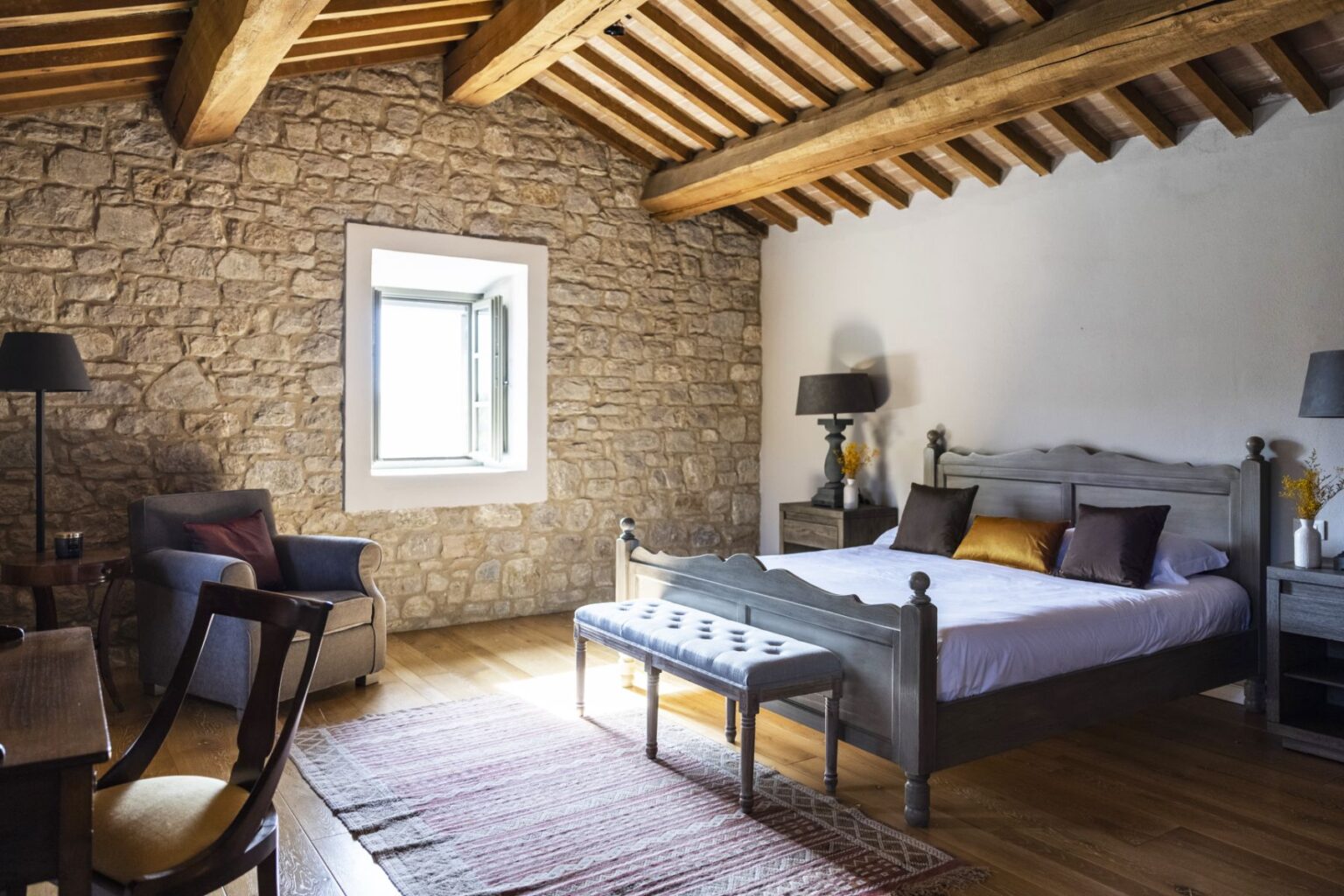 Cozy bedroom featuring a stone wall, wooden beams, and a comfortable bed with decorative pillows. The room includes a plush armchair, a wooden desk, and a stylish bench, illuminated by natural light from the window. | Only on SopranoVillas
