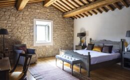 Cozy bedroom featuring a stone wall, wooden beams, and a comfortable bed with decorative pillows. The room includes a plush armchair, a wooden desk, and a stylish bench, illuminated by natural light from the window.