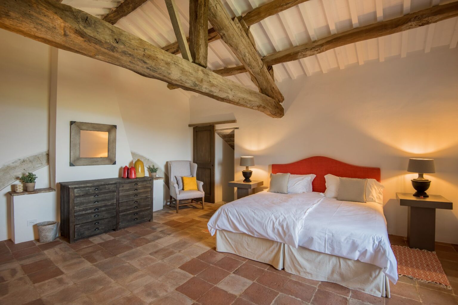 Spacious bedroom with wooden beams, featuring twin beds with white linens, a rustic wooden dresser, a cozy armchair, and warm lighting. The room has terracotta tile flooring and a minimalist design, creating a serene and inviting atmosphere. | Only on SopranoVillas