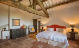 Spacious bedroom with wooden beams, featuring twin beds with white linens, a rustic wooden dresser, a cozy armchair, and warm lighting. The room has terracotta tile flooring and a minimalist design, creating a serene and inviting atmosphere.