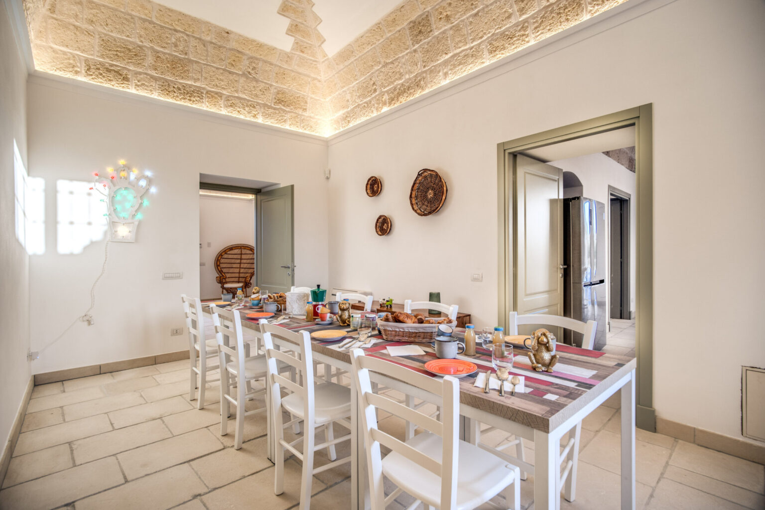 Spacious dining room featuring a long white table set for a meal, surrounded by white chairs, with decorative wall art and warm lighting, creating an inviting atmosphere for gatherings. | Only on SopranoVillas