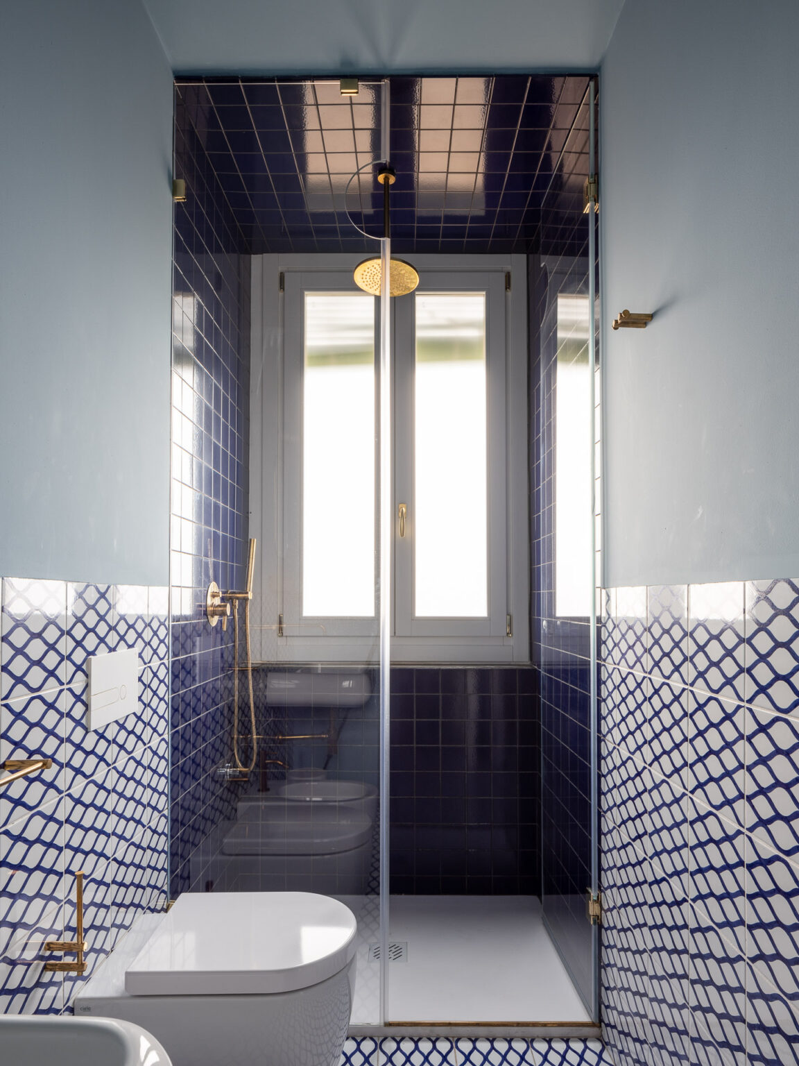 Modern bathroom featuring a glass shower enclosure with a gold shower head, blue tiled walls, and a stylish white toilet. Natural light streams through the window, highlighting the contemporary design and elegant fixtures. | Only on SopranoVillas