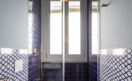 Modern bathroom featuring a glass shower enclosure with a gold shower head, blue tiled walls, and a stylish white toilet. Natural light streams through the window, highlighting the contemporary design and elegant fixtures.