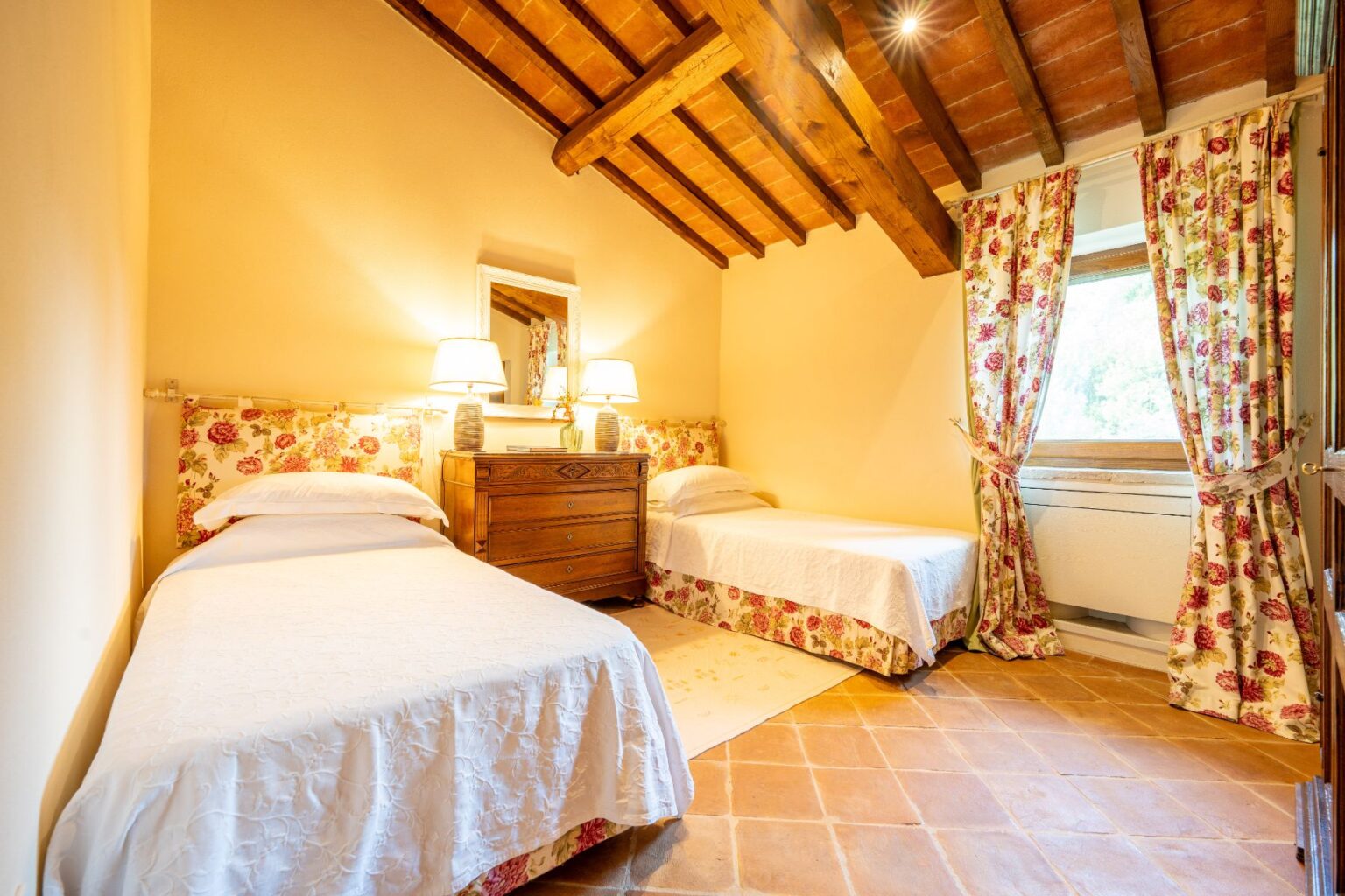Cozy bedroom featuring two twin beds with white bedding, a wooden dresser, and floral curtains. The room has a warm ambiance with exposed wooden beams and natural light from a window. Ideal for relaxing stays in a charming setting. | Only on SopranoVillas
