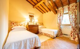 Cozy bedroom featuring two twin beds with white bedding, a wooden dresser, and floral curtains. The room has a warm ambiance with exposed wooden beams and natural light from a window. Ideal for relaxing stays in a charming setting.