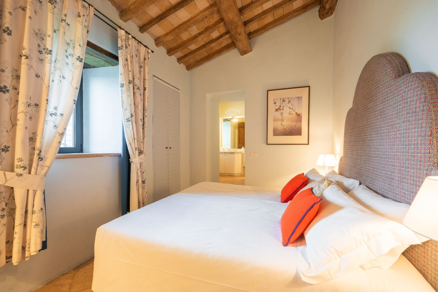 Cozy bedroom featuring a king-sized bed with decorative orange pillows, wooden beam ceiling, and floral curtains. The room includes a window with natural light, a wardrobe, and a framed artwork on the wall, creating a warm and inviting atmosphere. Ideal for a relaxing getaway. | Only on SopranoVillas