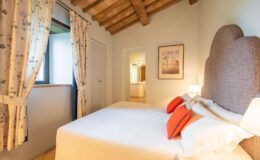 Cozy bedroom featuring a king-sized bed with decorative orange pillows, wooden beam ceiling, and floral curtains. The room includes a window with natural light, a wardrobe, and a framed artwork on the wall, creating a warm and inviting atmosphere. Ideal for a relaxing getaway.