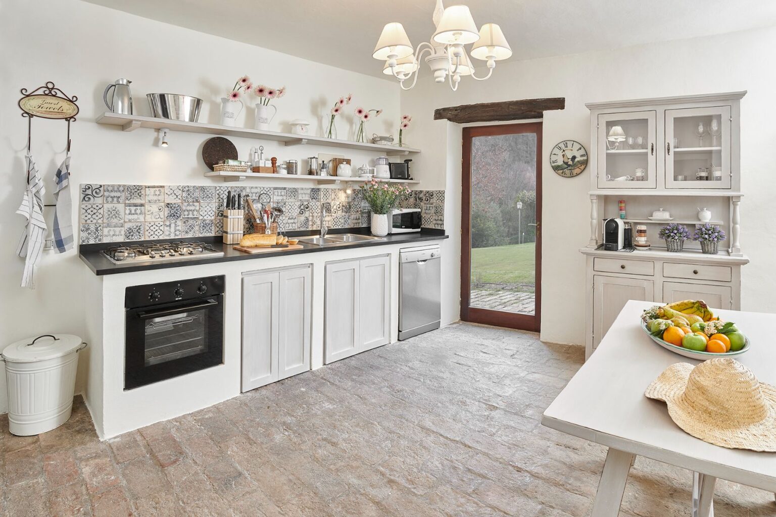 Bright and airy kitchen featuring white cabinets, a modern gas stove, and decorative tiles. Open shelving displays kitchenware and flowers, while a dining table with fresh fruit and a straw hat adds a cozy touch. Natural light streams in from a glass door leading to an outdoor space. | Only on SopranoVillas