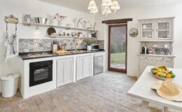 Bright and airy kitchen featuring white cabinets, a modern gas stove, and decorative tiles. Open shelving displays kitchenware and flowers, while a dining table with fresh fruit and a straw hat adds a cozy touch. Natural light streams in from a glass door leading to an outdoor space.